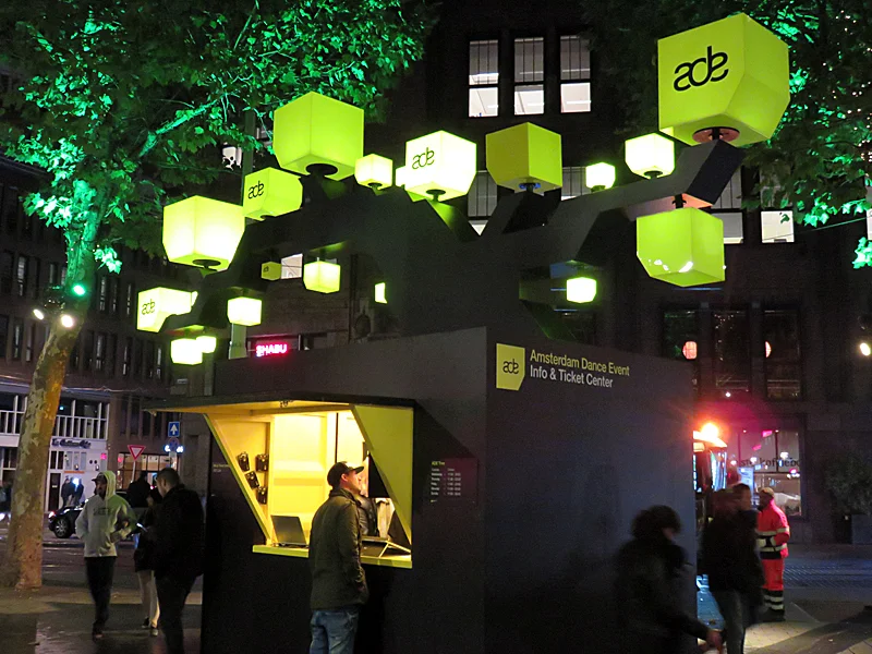 Yellow ADE cubes illuminate an information and ticket kiosk beneath the trees on Amsterdam’s Rembrandtplein at night.